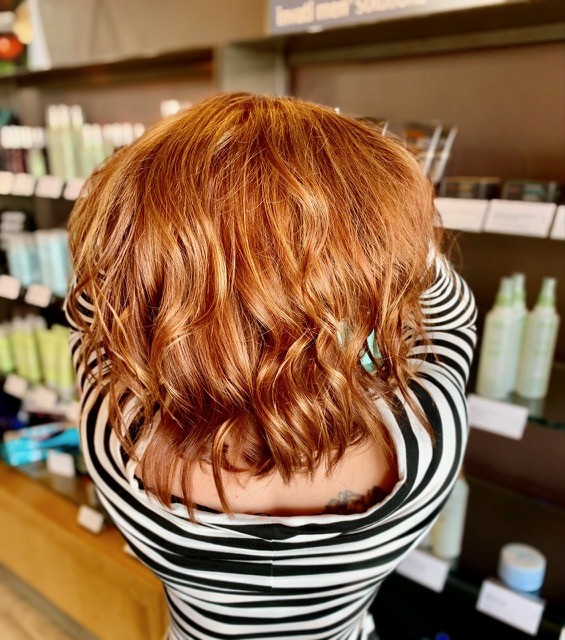 Salon Indigo in Fraser, MI A person with wavy, shoulder-length auburn Hair Color stands facing away, wearing a striped top, in front of shelves with hair care and Skin Care products at an Aveda Salon. Salon Indigo in Fraser, MI