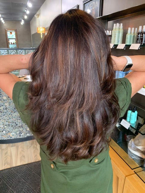 Salon Indigo in Fraser, MI Person with long, layered brown hair stands in an Aveda Salon, facing away from the camera with hands behind head, surrounded by shelves lined with premium hair products. Salon Indigo in Fraser, MI