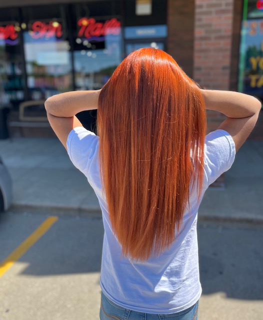 Salon Indigo in Fraser, MI Person with long, straight, vibrant red hair stands outdoors facing away from the camera, wearing a white t-shirt in front of a storefront—showcasing gorgeous locks that any hair stylist would admire. Salon Indigo in Fraser, MI