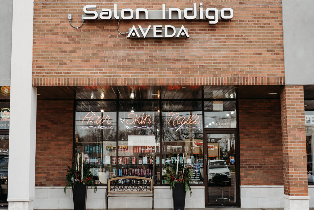 Salon Indigo in Fraser, MI Exterior of Salon Indigo AVEDA, an Aveda Salon, with large windows displaying "Hair Skin Nails" in neon signs; shelves with hair products, skin care items, and potted plants are visible inside. Salon Indigo in Fraser, MI