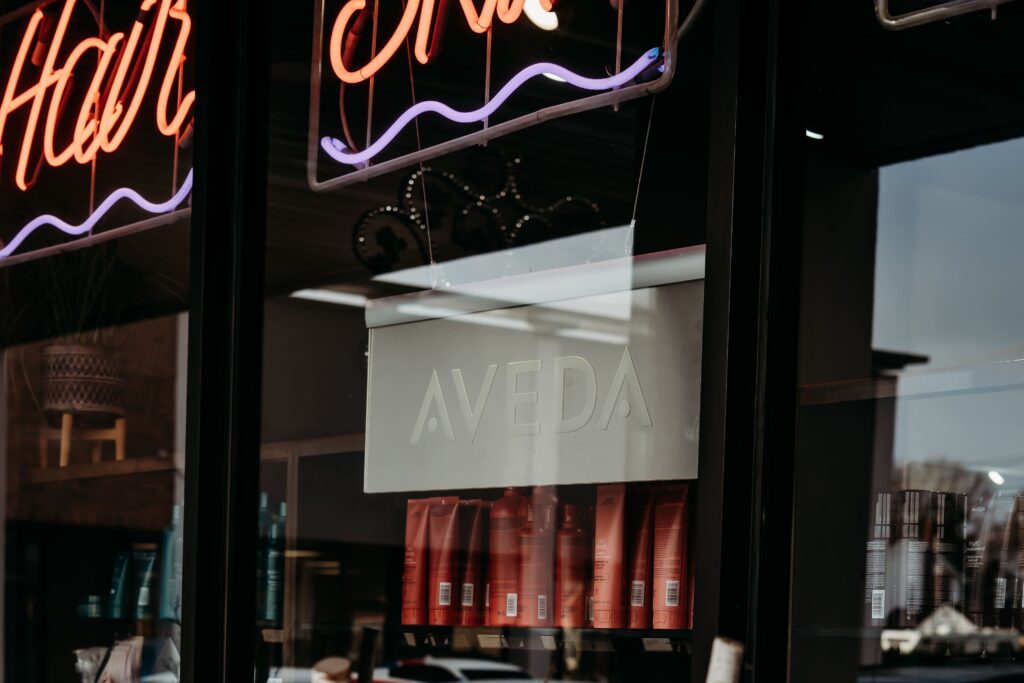 Salon Indigo in Fraser, MI Storefront window with a neon sign and a reflective glass displaying a white "Aveda" sign above shelves with red Aveda Salon hair products. Salon Indigo in Fraser, MI
