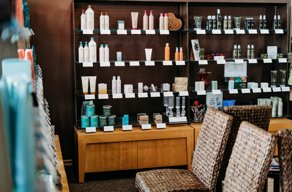 Salon Indigo in Fraser, MI Shelves display various skincare and haircare products in bottles and tubes, with price tags, next to a seating area with woven chairs in an Aveda Salon, creating a welcoming space for clients awaiting their hair stylist. Salon Indigo in Fraser, MI