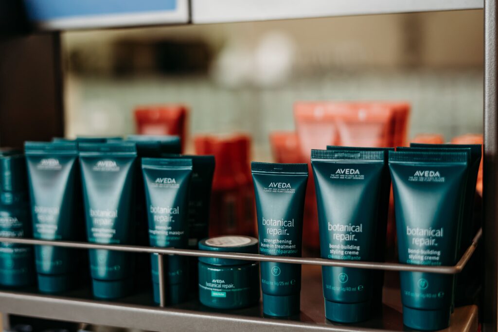 Salon Indigo in Fraser, MI Several teal-colored Aveda Botanical Repair hair care products are displayed on a metal shelf, with other red bottles and Skin Care items blurred in the background. Salon Indigo in Fraser, MI