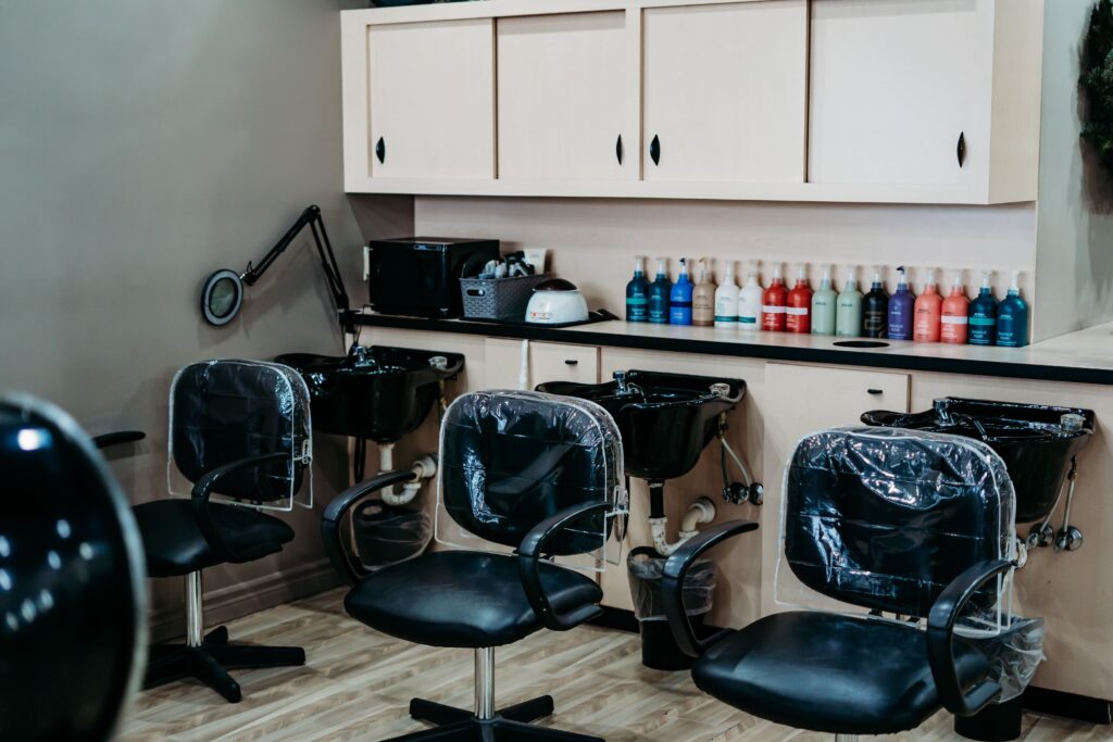 Salon Indigo in Fraser, MI Three salon chairs with plastic covers are positioned in front of washing sinks, while shelves above display hair products, creating a modern salon setting perfect for a professional hair stylist or manicure pedicure services. Salon Indigo in Fraser, MI
