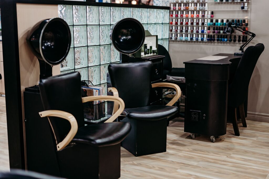 Salon Indigo in Fraser, MI A chic Aveda Salon interior featuring two black styling chairs, hooded hair dryers, a workstation, and shelves stocked with nail polish bottles—perfect for your next manicure pedicure appointment. Salon Indigo in Fraser, MI