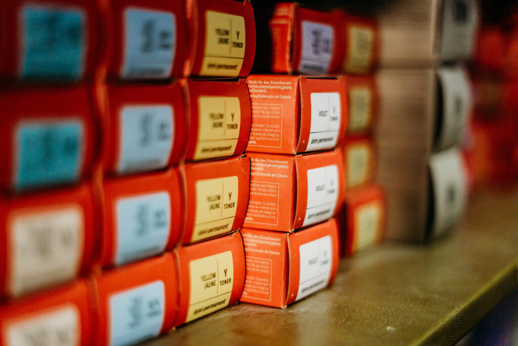 Salon Indigo in Fraser, MI Stacks of small orange and yellow hair color boxes with color labels are arranged on a shelf, viewed from a close-up angle. Salon Indigo in Fraser, MI